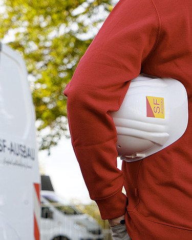 Person mit SF-Ausbau-Schutzhelm vor Firmenfahrzeug im Außeneinsatz.